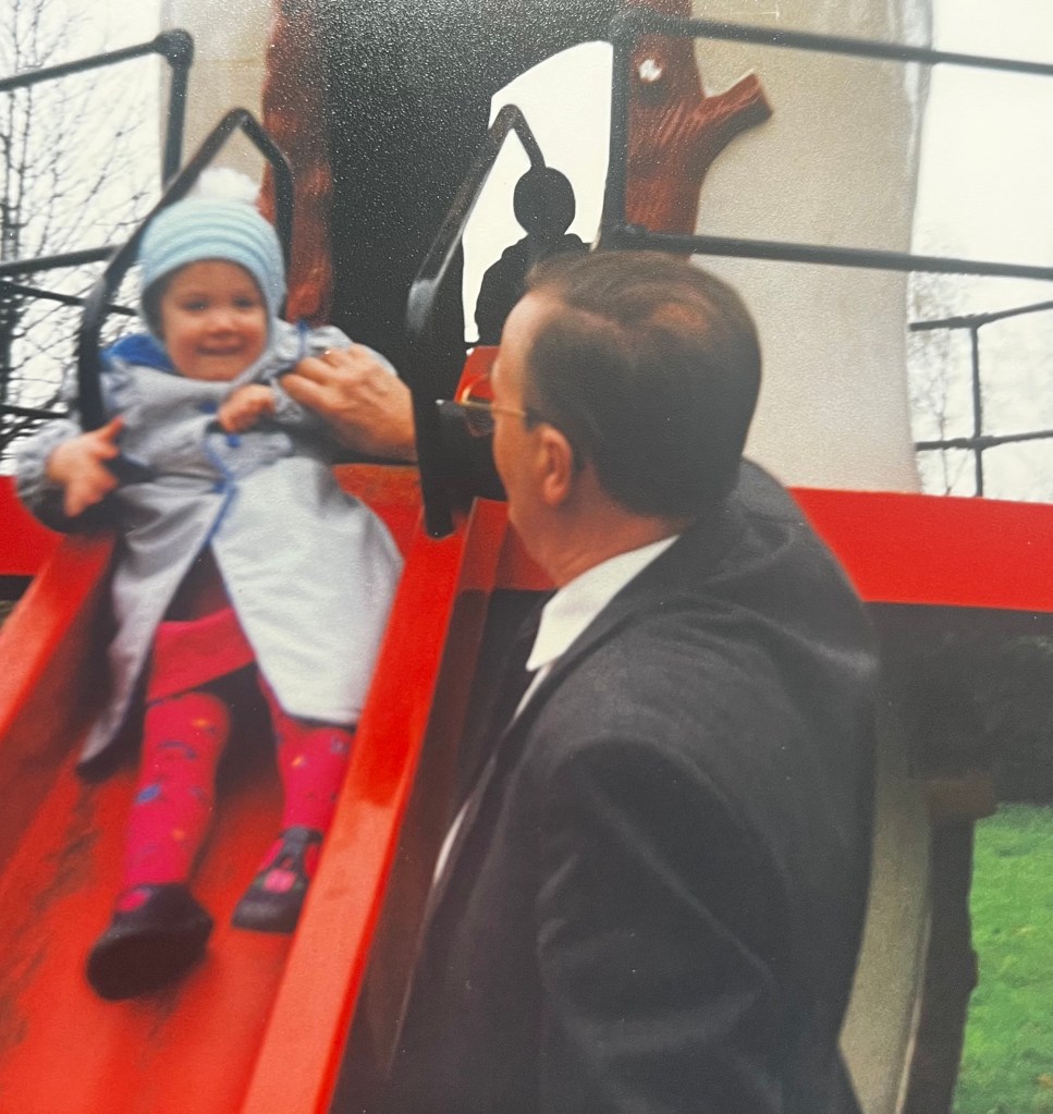 A photo of Rachael with her Grandad when she was a child. He is helping her down a slide in a play area.